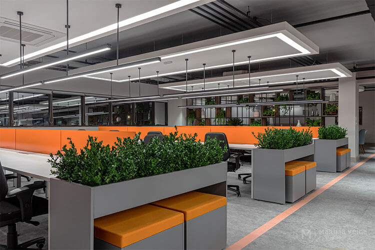 cores na decoração em escritório com estilo industrial, cor predominante cinza com detalhes em laranja na divisão entre as mesas e plantas nas laterais