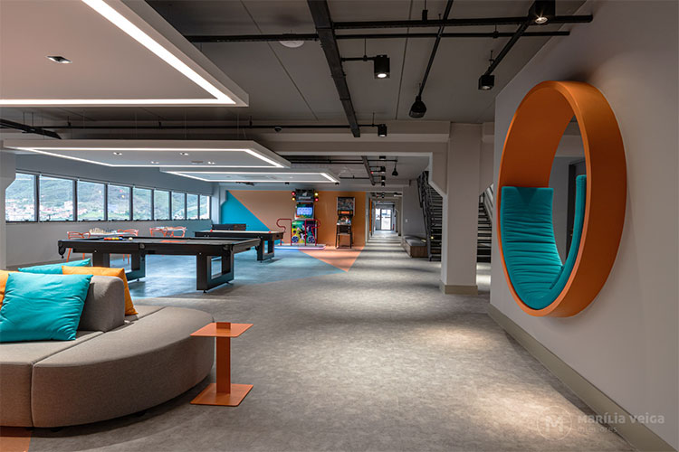 cores na decoração de lounge de empresa, ambiente amplo com sofá, mesa de bilhar e jogos. Cor predominante cinza com detalhes em laranja e azul turquesa