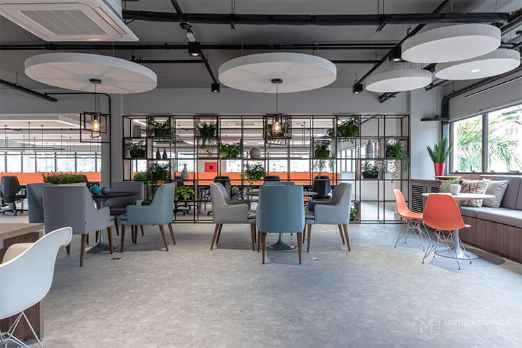 cores na decoração de um lounge de um escritório com mesas redondas e quatro cadeiras estofadas em volta, em estilo industrial com tons cinzas e detalhes em laranja