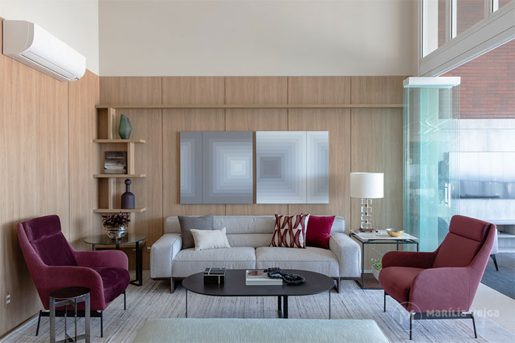 cores na decoração em sala de estar com painel de madeira em duas paredes, sofá cinza claro com almofadas cinzas e bordô, poltronas bordô (uma de cada lado), mesa de centro preta e porta de vidro aberta para integração com a varanda a direita