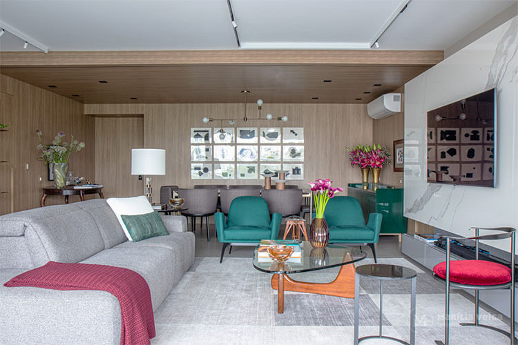 cores na decoração em sala de estar integrada com sala de jantar, painel de madeira e mesa de jantar com 8 cadeiras estofadas em cinza ao fundo, sofá cinza claro a esquerda, duas poltronas verdes a frente, tv em painel branco a direita e mesa de centro de vidro com pés de madeira