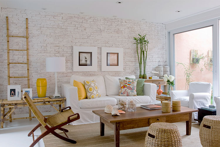 cores na decoração de uma sala de estar com parede do fundo de tijolo branco, sofá e poltrona brancos, móveis em madeira a bambu e detalhes da decoração como almofadas e vasos em tons de amarelo, verde e laranja