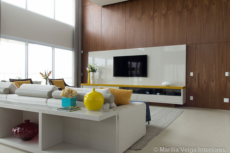 Painel branco atrás da tv, sobre painel maior de madeira escura, rack branco com tampo em amarelo, sofá branco com aparador atrás e detalhes em amarelo na decoração como poltronas, almofadas e vasos