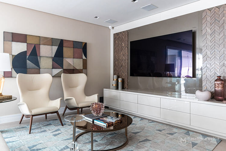 Living com tv em painel cinza escuro com detalhes geométricos nas laterais, rack branco com vasos de decoração em cima, quadro colorido e geométrico na parede ao lado com duas poltronas of white na frente e mesa de centro redonda e escura sobre tapete claro com desenho de triângulos