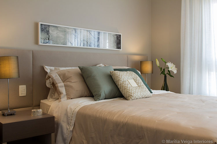 Cama com painel estofado em tom similar ao da parede, quadro retangular comprido acima, um abajur de cada lado, roupa de cama nude e almofadas verdes