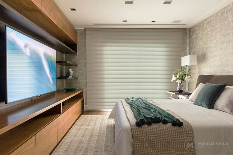 suítes de casal com cores claras e detalhes de decoração em verde. Do lado oposto da cama tem um móvel grande em madeira com armários e a tv posicionada no centro