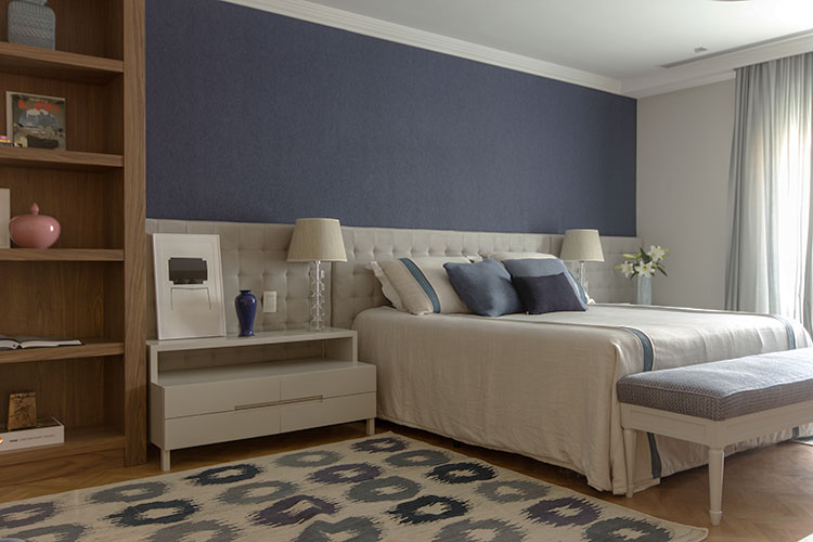 suítes de casal com detalhes da decoração em azul na parede atrás da cama, almofadas e estampa do tapete, roupa de cama clara, painel estofado, banco de apoio em frente a cama e estante de madeira ao lado
