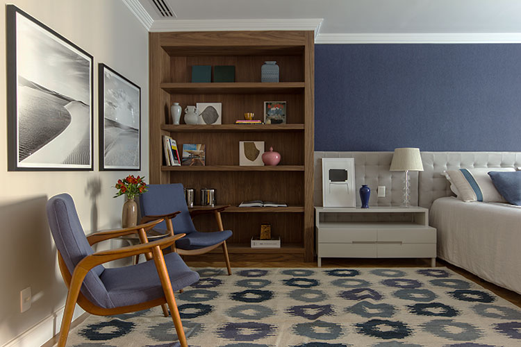 suítes de casal com detalhes da decoração em azul na parede atrás da cama, almofadas, estampa do tapete e poltronas encostadas na parede lateral, roupa de cama clara, painel estofado, mesa de cabeceira clara ao lado da cama e estante de madeira ao lado