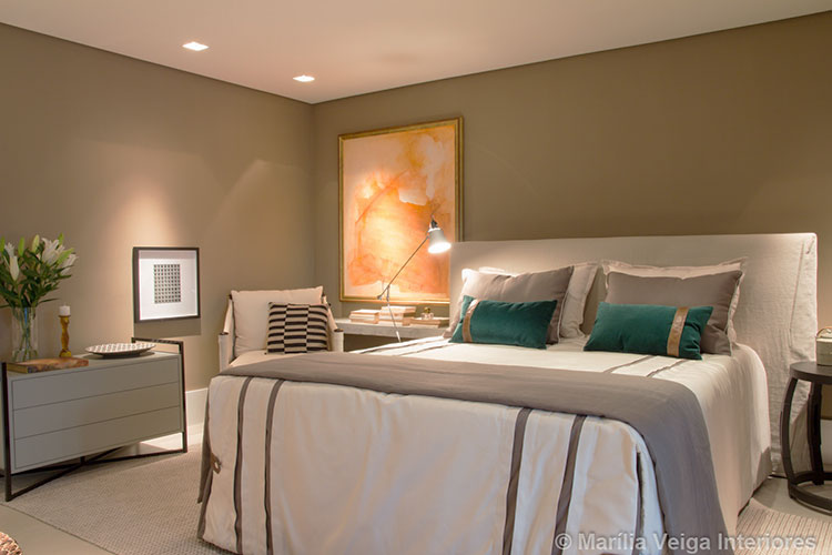 mesma suítes de casal que a anterior em outro ângulo, em tons nude mais escuros com quadros de decoração, poltrona e luminária ao lado da cama