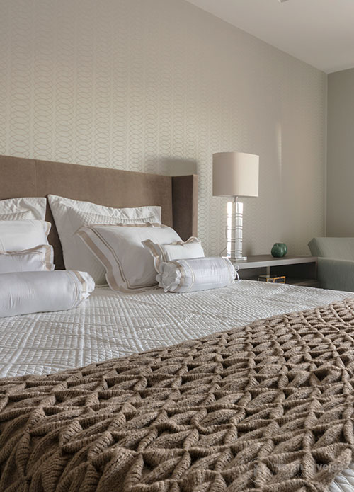 suítes de casal vista da cama com lençol claro e colcha marrom, parede com textura ao fundo