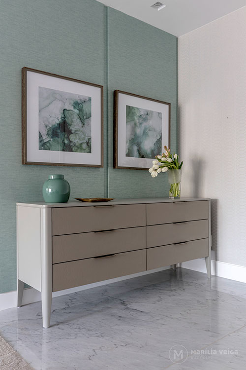 Detalhe de aparador em tom nude em um canto da suíte, parede verde claro atrás, dois quadros quadrados e vasos em cima
