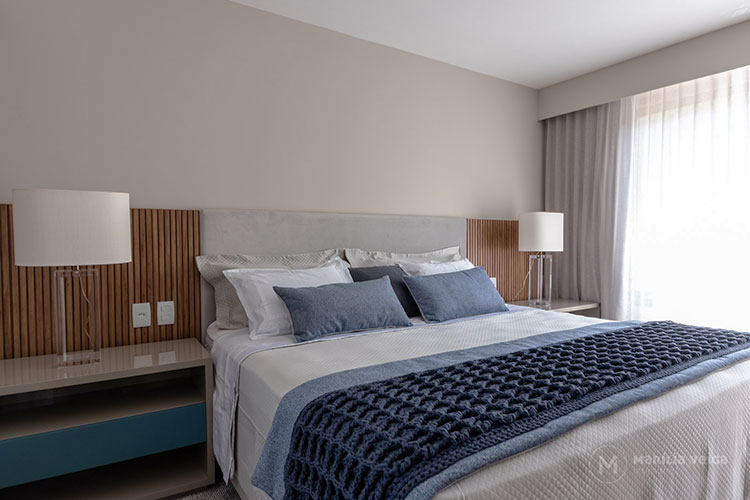 suítes de casal com painel estofado atrás da cama completado por um painel de madeira nas laterais, mesas de cabeceira com abajur, roupa de cama clara e almofada e manta em tons azuis
