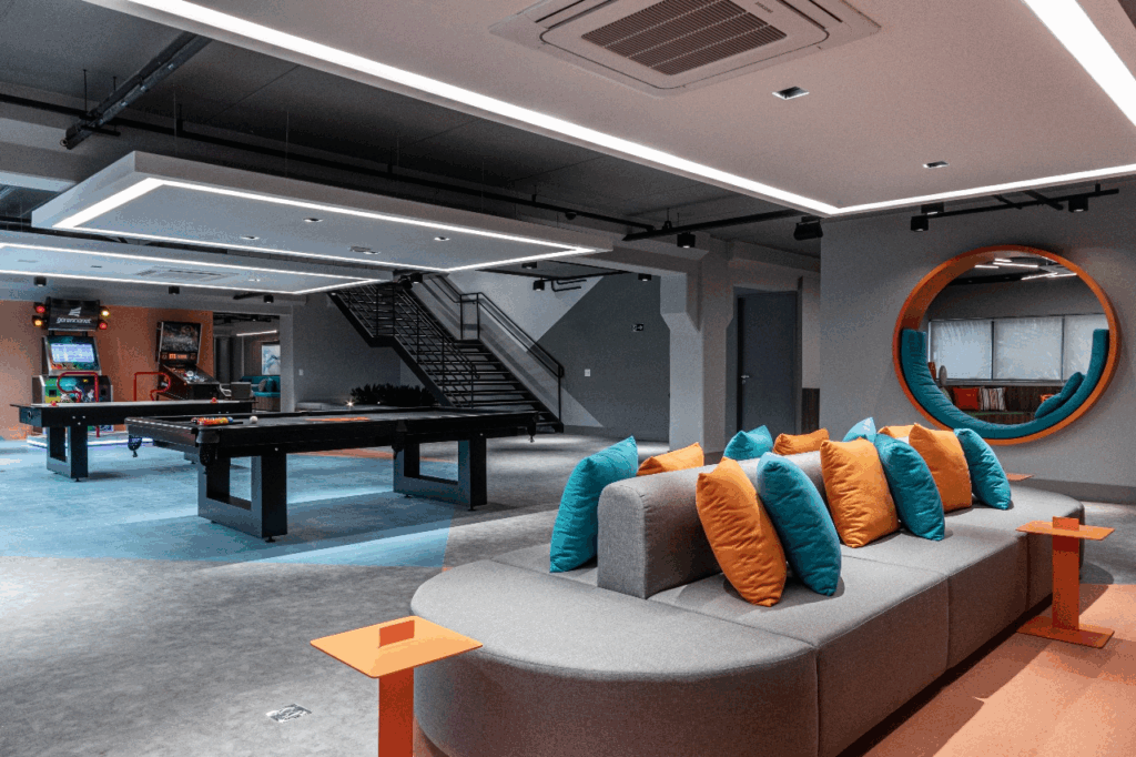 Vista ampla da área de descompressão da sede do Efí Bank — parte do projeto de escritório corporativo — com mesas de bilhar, máquinas de arcade, sofá curvo com almofadas em laranja e azul e iluminação linear no forro aparente. A paleta da marca organiza o espaço e cria pontos-cenário fotogênicos. O layout aberto favorece circulação e encontros rápidos sem disputar salas. Esta composição ajudou a transformar as duas lajes em um escritório instagramável que as pessoas gostam de usar no dia a dia, impulsionando convivência e colaboração.