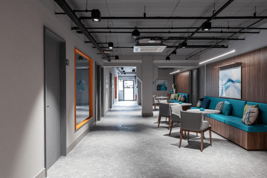 A imagem mostra como os corredores do Efí Bank foram incorporados ao projeto de escritório corporativo: à direita, marcenaria em madeira com bancos estofados azuis e mesas de apoio cria áreas para conversas rápidas. À esquerda, a moldura laranja marca o percurso e reforça a identidade visual. O forro aparente com trilhos e luminárias lineares garante luz homogênea, enquanto spots destacam as zonas de uso. O resultado é mais oferta de lugares de encontro sem ampliar a área construída — uma solução que acelera a colaboração no dia a dia.