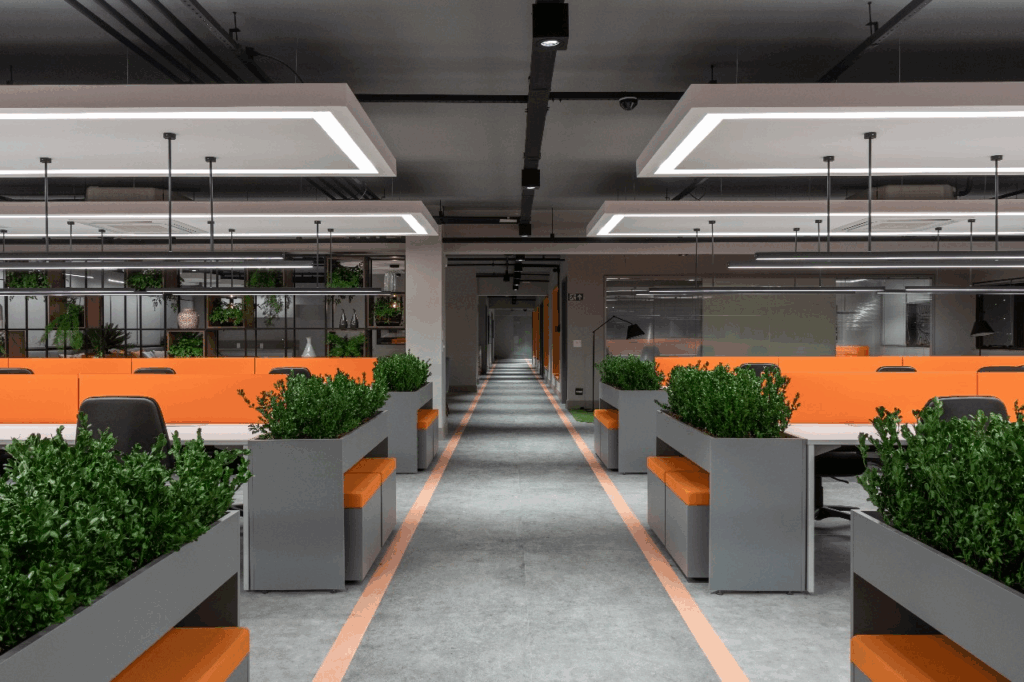 Vista do open space do Efí Bank com estações de trabalho divididas por jardineiras e bancos estofados em laranja. As luminárias lineares suspensas distribuem a luz de forma homogênea, reduzindo cansaço visual. A faixa no piso guia a circulação e ajuda na leitura do layout. O conjunto de materiais, verde e cores institucionais traduz o design de interiores corporativo em benefício prático: foco nas mesas, encontros curtos nos bancos laterais e circulação desobstruída — três pilares que sustentam produtividade ao longo do dia.