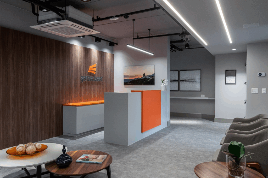 Recepção da sede do Efí Bank com painel em madeira e balcão cinza com destaque laranja, integrando identidade visual logo na entrada. As luminárias lineares no forro aparente criam iluminação uniforme, ideal para fotos e vídeos, enquanto a composição limpa facilita o fluxo de visitantes. Mesas de apoio e poltronas garantem conforto sem ruído visual. Este enquadramento funciona como cartão de visita do projeto de escritório corporativo: comunica quem a empresa é e oferece um cenário consistente para registros institucionais e de recrutamento.