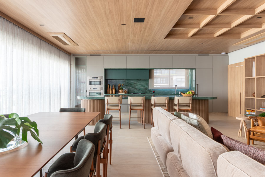 Integração total da parte social: cozinha aberta ao fundo com pedra verde, bancada com banquetas, sala de estar com sofá bege abraçador, sala de jantar com mesa de madeira, estante de marcenaria que abraça toda a parede. Apartamento Jardim Paulista, Marília Veiga.